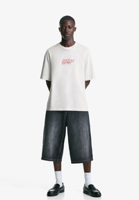 Tricou alb din bumbac cu text roșu "HEART", asortat cu pantaloni scurți din denim închis. Modelul poartă pantofi negri și șosete albe. Fundal de studio.