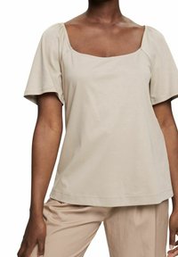 Femme portant un haut beige à manches courtes avec un col carré et un pantalon beige assorti sur fond blanc.