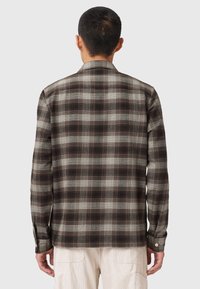 Man with short hair wearing a brown and gray plaid long-sleeve shirt and light-colored pants, shown from the back against a plain background.