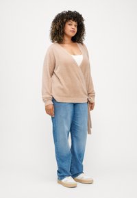 Μπεζ ζακέτα με απαλή υφή και μακριά μανίκια, δεμένη στη μέση. Φοριέται πάνω από ένα λευκό μπλουζάκι με ανοιχτό μπλε τζιν κατευθείαν πόδια και λευκά sneakers.