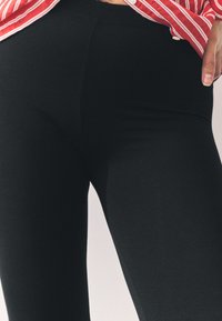 Svarte leggings lavet af strækstof, med en glat tekstur og et tætsiddende design, delvist med en stribet skjorte synlig ved taljen.