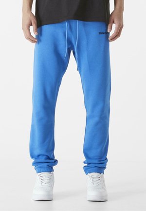 Blaue Sweatpants aus weichem Stoff, mit einem elastischen Bund und Bündchen, versehen mit einem schwarzen Logo, das nahe der Taille gedruckt ist. Getragen mit weißen Sneakers.