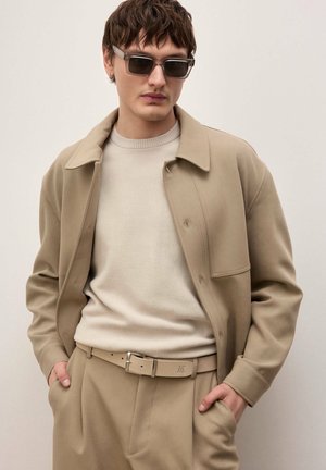 Noor mees, kes kannab beežikat jopet, kreemjat soonikut kampsunit, beeže pükse sobiva vööga ja ristkülikukujulisi päikeseprille, käed taskutes.