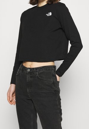 Kvinde iført en sort cropped langærmet top med The North Face-logo og sorte højttaljede jeans, den ene hånd i lommen.