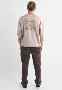 Sweatshirt beige avec un design floral au dos, avec des accents gris et jaune. Associé à un pantalon marron et des baskets noires.
