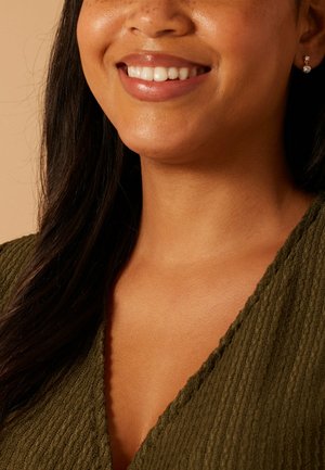 Lachende vrouw met lang, donker haar, die een groen, gestructureerd topje en een kleine ronde diamantoorbel draagt tegen een beige achtergrond.