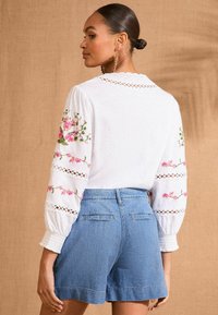 Bijela bluzica s cvjetnim vezom u ružičastoj i zelenoj boji, napuhane rukave s čipkastim detaljima, uparena s plavim trapericama. Jednostavan i teksturiran dizajn.