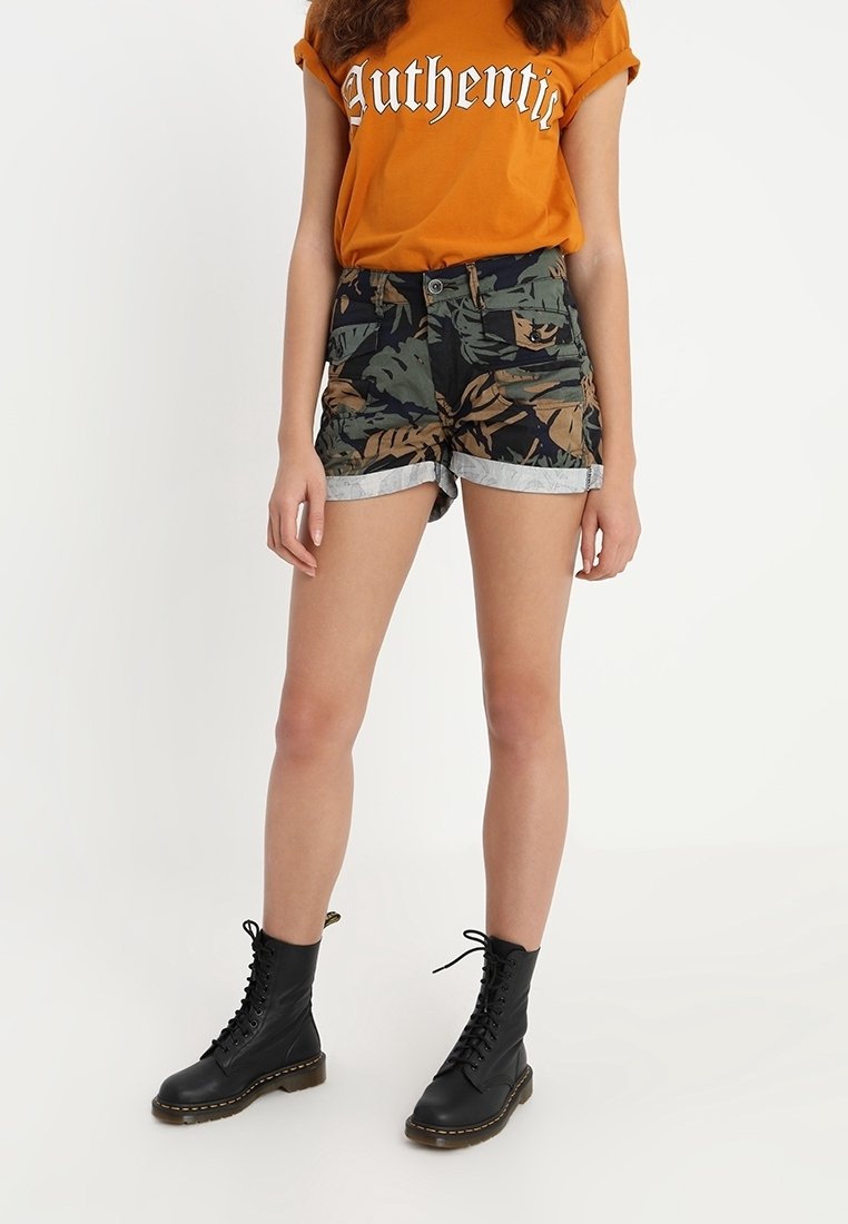 Oranžové tričko s textem "Autentické"; kraťasy s maskáčovým vzorem a ohrnutými lemy; černé šněrovací boty. Neformální outfit s kombinací různých textur.