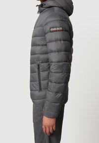 Man wearing a gray quilted puffer jacket with a hood, zippered side pocket, and a Napapijri Geographic logo on the upper sleeve.