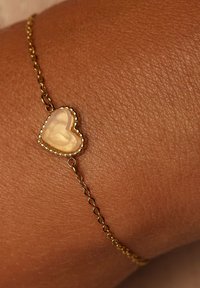 Goldene Kettenarmband mit einem herzförmigen, schimmernden Anhänger mit strukturierter Oberfläche und Perlenrand, der auf einem Handgelenk ruht.
