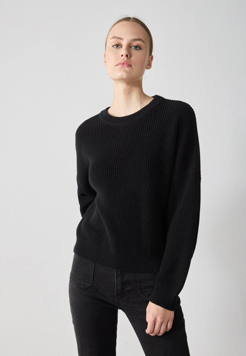 Svart strikket genser med rund hals og lange ermer. Har ribbestrikket tekstur og cropped passform, kombinert med svarte jeans.