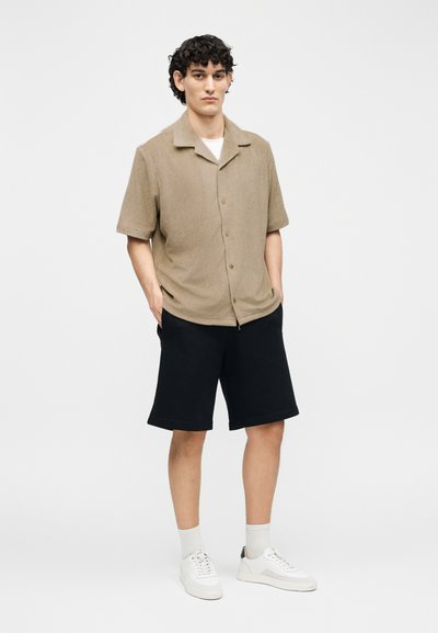 Νέος άνδρας με σγουρά μαλλιά που φοράει μπεζ πουκάμισο με κοντά μανίκια και κουμπιά, μαύρο σορτς, άσπρες κάλτσες και άσπρα αθλητικά παπούτσια, στέκεται με τα χέρια στις τσέπες.