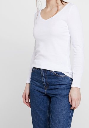 Vrouw draagt een nauwsluitend wit shirt met lange mouwen en een V-hals, gecombineerd met een hooggetailleerde blauwe spijkerbroek, tegen een effen achtergrond.