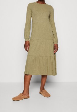 Kvinde iført en olivengrøn, langærmet kjole med lag i midi-længde og brune, bløde slip-on sko, stående mod en ensfarvet baggrund.