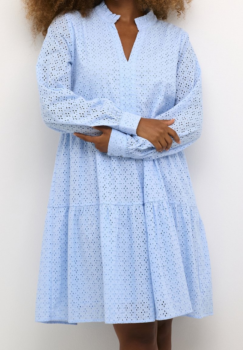 Femme aux cheveux bouclés portant une robe bleu clair à manches longues avec une jupe froncée, debout les bras croisés.