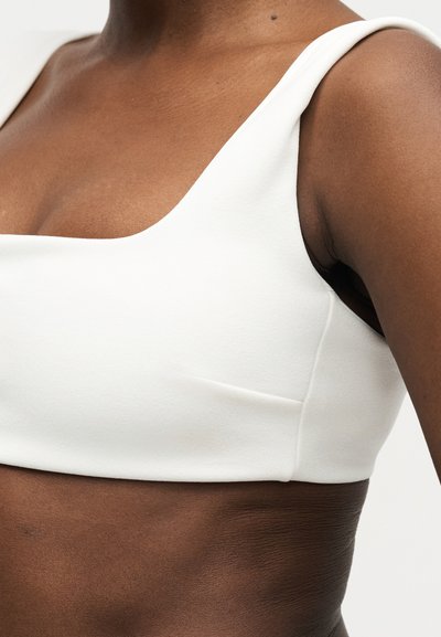 Πρόσωπο με σκουρόχρωμο δέρμα που φοράει λευκό αμάνικο crop top, εμφανίζοντας τον ώμο και το πάνω μέρος του στήθους με λείο ύφασμα και εφαρμοστή σχεδίαση.