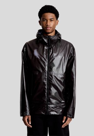 Ung mand iført en blank sort hættejakke med lynlås foran og lommer, stående mod en lys grå baggrund.