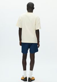 T-shirt en coton crème à manches courtes, associé à un short bleu marine. Le modèle porte des chaussettes blanches et des chaussures jaune-noir, vu de dos.