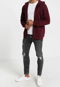 Sudadera burdeos con cremallera y bolsillo frontal, llevada sobre una camiseta blanca. Combinada con jeans negros desgastados y zapatillas blancas con detalles rojos.
