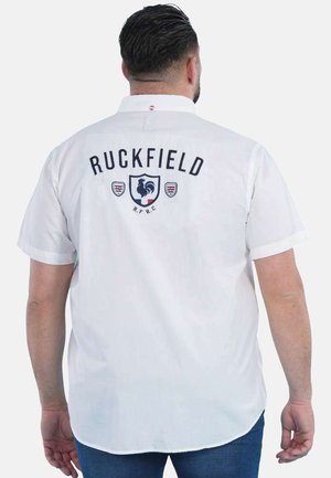 Homme portant une chemise blanche à manches courtes avec "Ruckfield" et un emblème de coq dans le dos, associée à un jean bleu.
