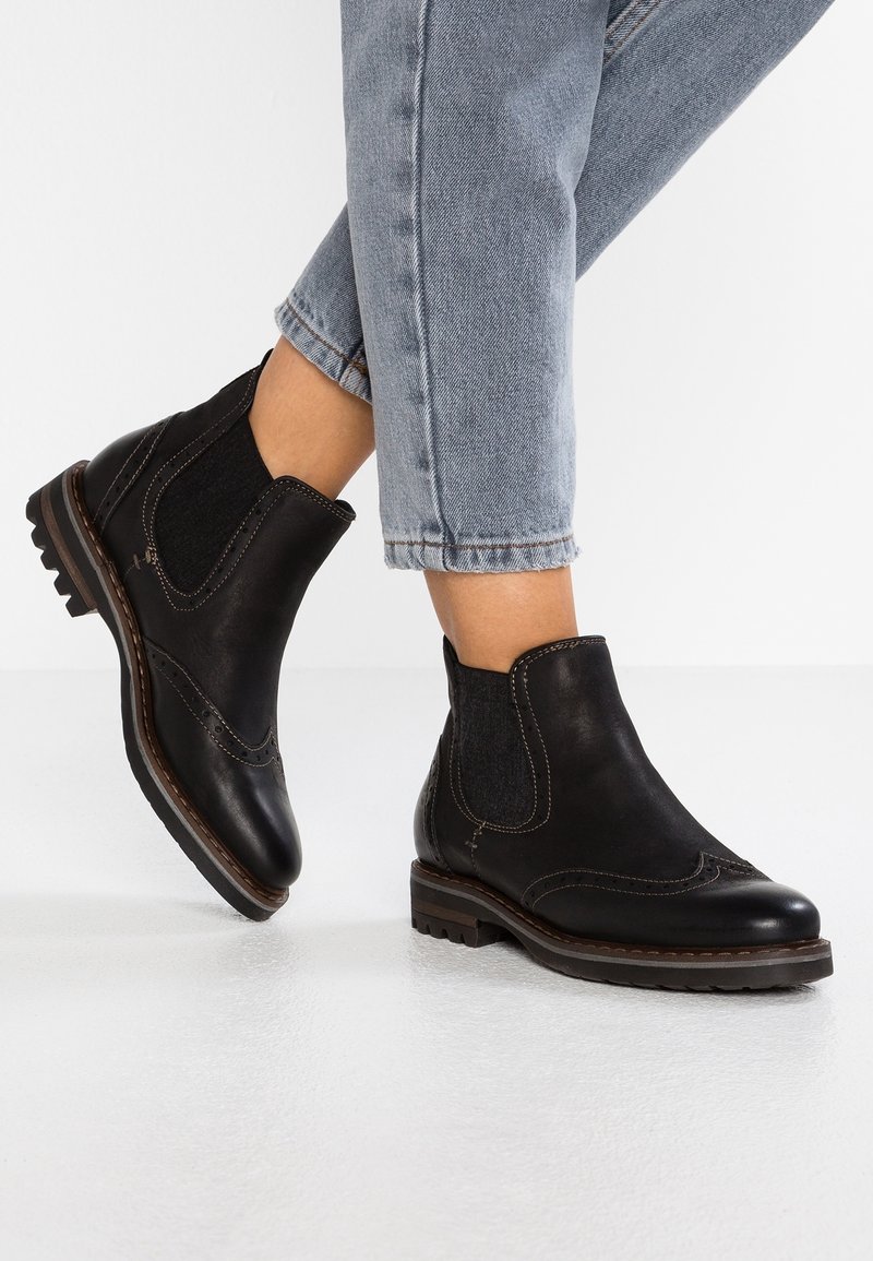 Černé kotníkové kožené boty s elastickými bočními panely, brogue detaily a texturovanými podrážkami, kombinované s kratšími modrými džínami.