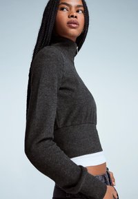 Maglione corto grigio scuro con collo alto e maniche lunghe, caratterizzato da una texture a coste e un design aderente. Indossato sopra una maglietta bianca.