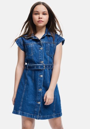 Ung kvinde iført en knappet, ærmeløs denimkjole med bælte og krave, der står foran en ensfarvet lys baggrund.