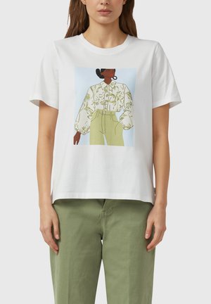 Frau trägt weißes T-Shirt mit Grafik einer dunkelhäutigen Frau in Blumenhemd und hellgrüner Hose, kombiniert mit olivgrünen Hosen.