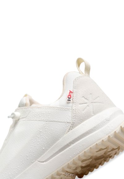 Bílé sportovní boty s texturovanou gumovou podrážkou, voděodolným svrškem a minimalistickým designem. Obsahují malou červenou visačku a kapky vody.