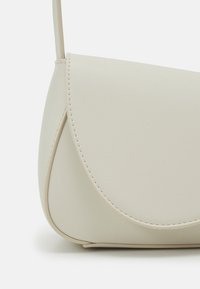 Sac bandoulière en cuir couleur crème avec rabat courbé et coutures visibles sur un fond uni.