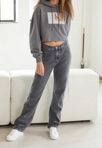 Osoba ubrana w szary skrócony hoodie Calvin Klein, ciemnoszare dżinsy z wysokim stanem i białe sneakersy stoi w pomieszczeniu obok białej sofy.