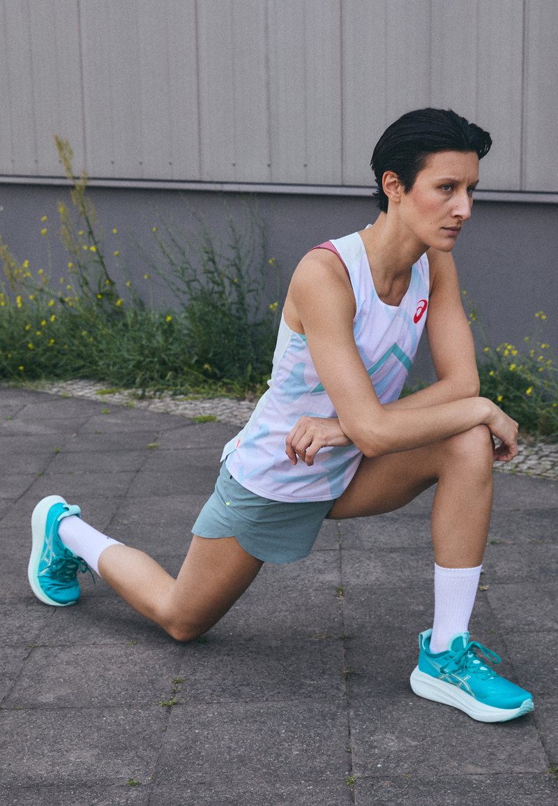 Sportowa koszulka na ramiączkach w jasnych pastelowych kolorach i geometrycznych wzorach, zestawiona z zielonymi szortami oraz turkusowymi butami do biegania; widoczne białe skarpetki sięgające do kostek.