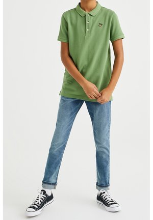 Zelené polo tričko se třemi knoflíky a malým vyšívaným logem, kombinované se světle modrými džínami a černými teniskami. Viditelné jsou bílé ponožky.