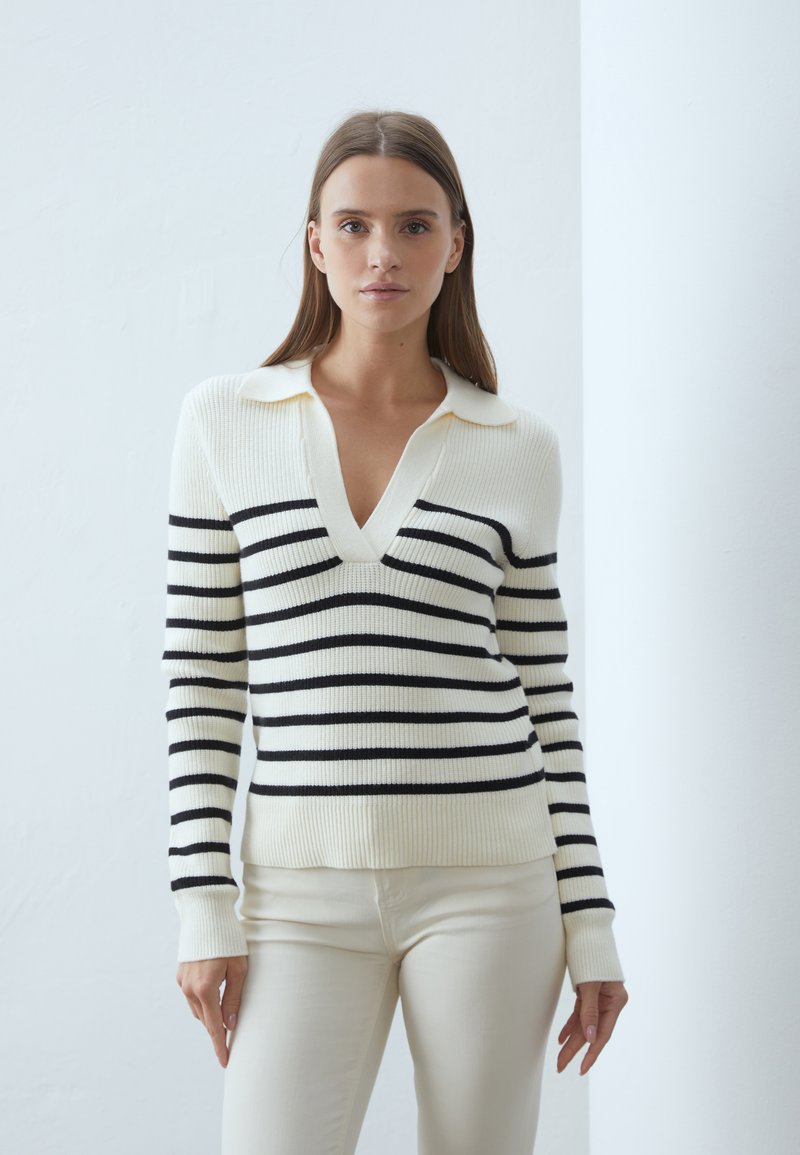 Pull tricoté noir et blanc à rayures avec un col en V et un col ; texture côtelée et design ajusté. Associé à un pantalon couleur crème.