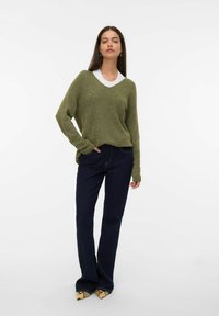 Vrouw in een olijfgroen oversized trui, met een witte blouse eronder, donkere spijkerbroek en schoenen met slangenprint met puntige neuzen, poseert met één hand in de zak.
