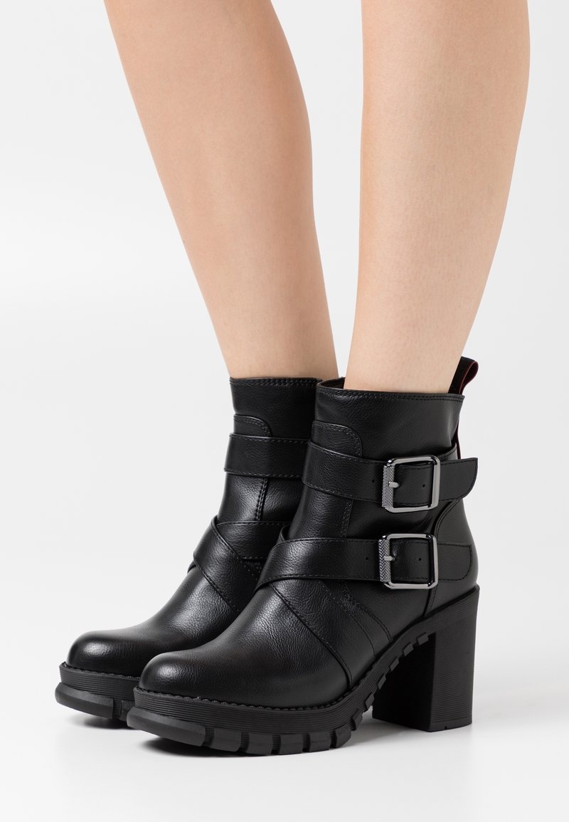 Botas de tobillo de cuero negro con dos correas decorativas y hebillas plateadas. Tacón alto grueso y suela de goma texturizada. Acabado suave.