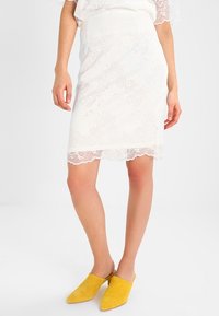 Weißer Spitzen-Bleistiftrock mit gewelltem Saum und floralen Mustern, kombiniert mit gelben Wildleder-Mules. Glatte Textur, figurbetonter Schnitt.