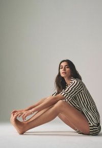 Gestreept kort mouwen shirt in zwart en wit, gecombineerd met shorts. Model zittend, met een gladde huid en lange benen. Minimalistische achtergrond.