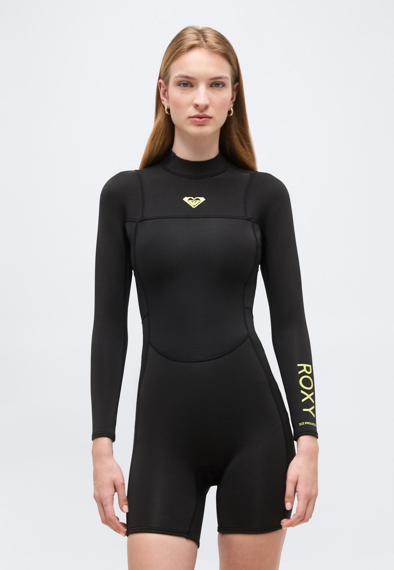 Femme portant une combinaison de plongée noire à manches longues avec un logo Roxy jaune sur la poitrine et la manche, debout devant un fond clair uni.