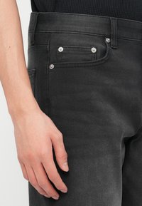 Κοντινό πλάνο ενός χεριού που ακουμπά δίπλα σε τσέπη μαύρου τζιν παντελονιού με ασημένιες λεπτομέρειες σε βίδες και μαύρο υφασμάτινο πουκάμισο με υφή.