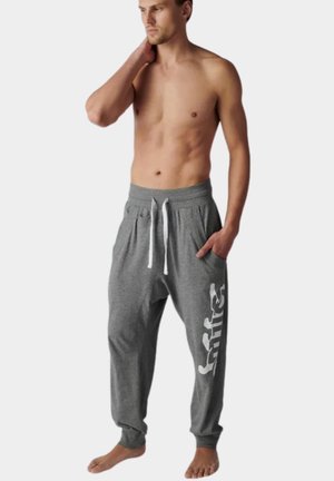 Mężczyzna bez koszulki stoi boso, jedną ręką w kieszeni, a drugą dotyka szyi, ubrany w szare spodnie dresowe z białymi sznurkami i logo na nogawce.