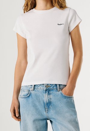 Femme portant un t-shirt blanc moulant à manches courtes avec un petit logo sur la poitrine et un jean en denim bleu clair taille haute, une main dans la poche.