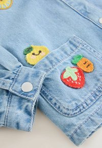Denimjackenärmel und -tasche mit bestickten, lächelnden Zitronen-, Orangen- und Erdbeer-Patches mit grünen Blättern und rosa Wangen.