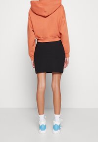 Orange kortärmad hoodie, svart ribbad kjol, vita och blå sportskor, synliga sömmar och en avslappnad passform. Enfärgad bakgrund.