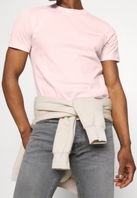 Camiseta rosa de manga corta con cuello redondo y detalle de logo, combinada con jeans grises y un sudadera color crema atada a la cintura.