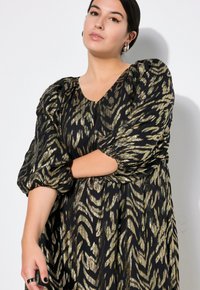 Musta mekko kultaisella ja vihreällä eläinkuosilla, jossa on pullotti hihat, syvä V-kaula-aukko ja virtaava siluetti. Sileä kangas, jossa on merkittävää kiiltoa.