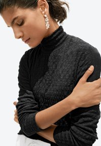 Frau in schwarzem, strukturiertem Rollkragenpullover mit kurzen Haaren trägt hängende geometrische Kristallohrringe und hat die Arme sanft vor der Brust verschränkt.