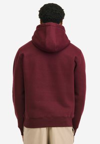 Tamnosmeđa majica s kapuljačom s velikom kapuljačom, teksturiranim materijalom, rebrastim manšetama i rubom. Pogled straga prikazuje glatki, neukrašeni dizajn.