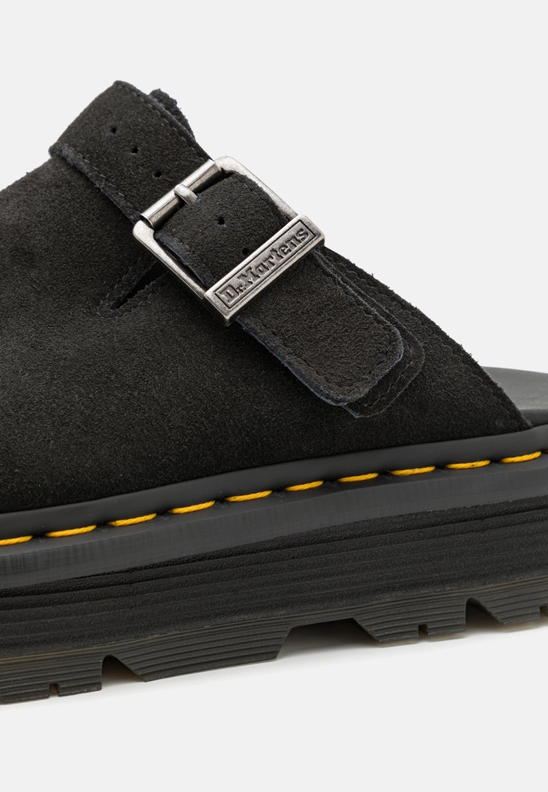 Schwarze Wildleder-Sandale mit einer metallischen Schnalle und Logo-Detail. Sie verfügt über eine dicke Sohle mit gelben Nähten und einem strukturierten Gummiprofil für besseren Halt.