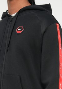 Fekete Nike kapucnis pulóvert viselő személy, piros és fehér logóval a mellen és piros Nike márkájú csíkkal az ujjon.